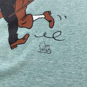 Pu&ograve; includere: T-shirt verde chiaro con un'illustrazione a cartoni animati di una figura in corsa in marrone e nero. Il design include una piccola grafica di un ragazzo e un cane, con la scritta "HERGE / MOULINSART" sotto. La maglietta ha un aspetto strutturato.