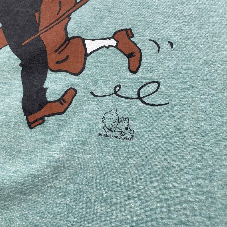 Pu&ograve; includere: T-shirt verde chiaro con un'illustrazione a cartoni animati di una figura in corsa in marrone e nero. Il design include una piccola grafica di un ragazzo e un cane, con la scritta "HERGE / MOULINSART" sotto. La maglietta ha un aspetto strutturato.