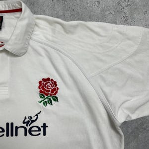 Pode incluir: Camisa de rugby branca com um emblema de rosa vermelha e folhas verdes no peito. A camisa tem gola e o texto "ellnet" em azul marinho. A camisa tem uma borda branca e vermelha.