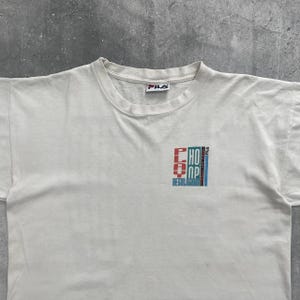 May include: White crew neck t-shirt with a FILA logo at the neckline. The shirt features a graphic design on the front that reads "CHOP" and "REGULATOR" in red, blue, and white. The shirt appears to be vintage.