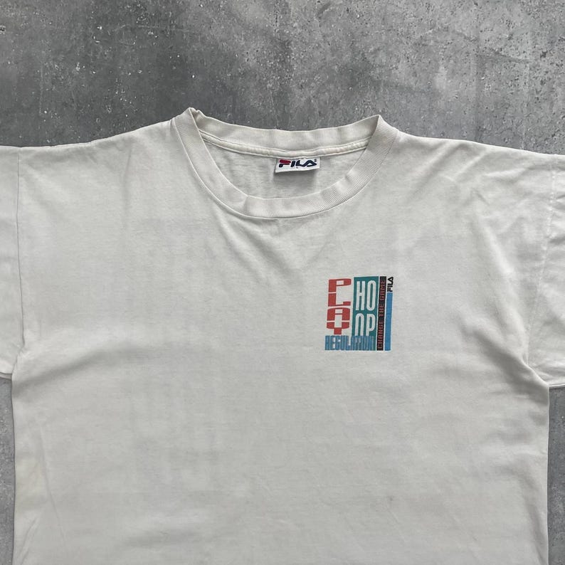 May include: White crew neck t-shirt with a FILA logo at the neckline. The shirt features a graphic design on the front that reads "CHOP" and "REGULATOR" in red, blue, and white. The shirt appears to be vintage.