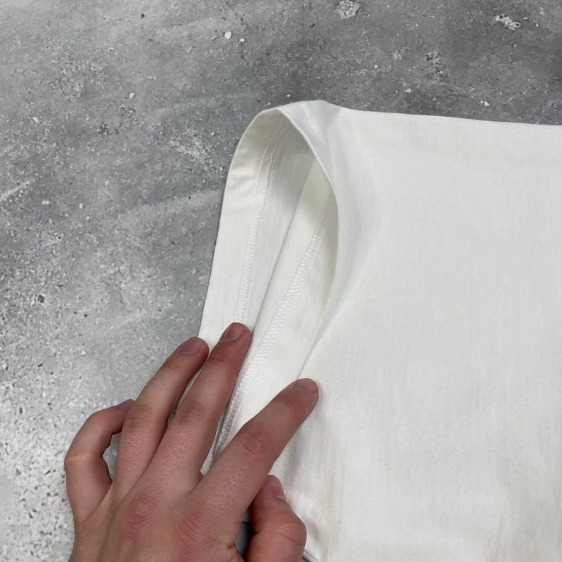 Peut inclure: Gros plan sur une manche de t-shirt blanche. La manche est faite d'un tissu lisse et opaque. Les coutures sont visibles et le tissu est pli&eacute;. L'arri&egrave;re-plan est une surface grise et textur&eacute;e.