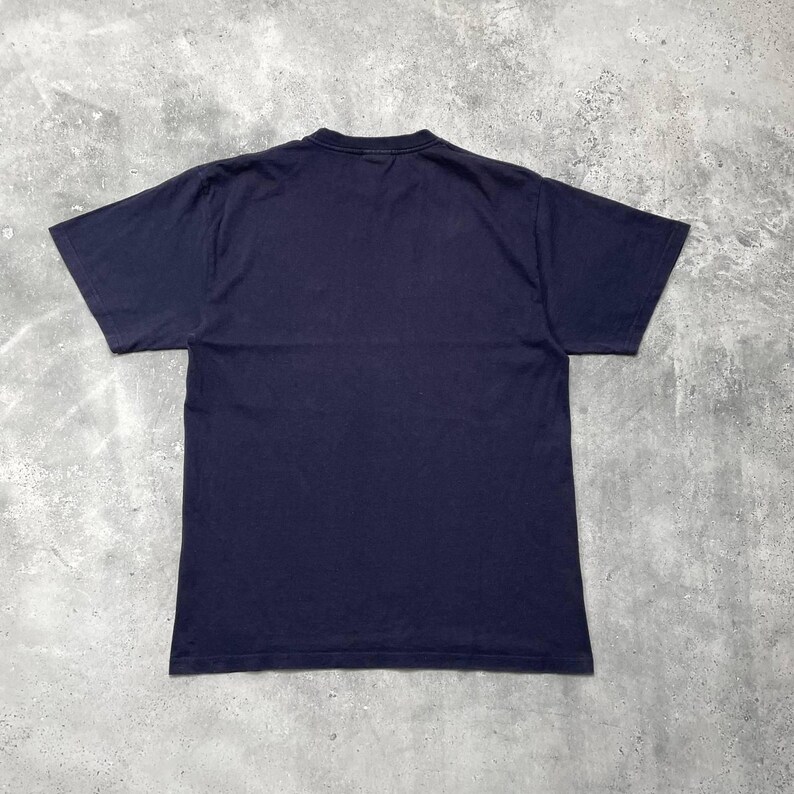 K&ouml;nnte beinhalten: Ein marineblaues T-Shirt, von hinten gesehen, liegt flach auf einer grauen Oberfl&auml;che. Das kurz&auml;rmelige Shirt besteht aus einem weichen Material und hat einen Rundhalsausschnitt. Das Shirt ist einfarbig, ohne sichtbare Grafiken oder Text.