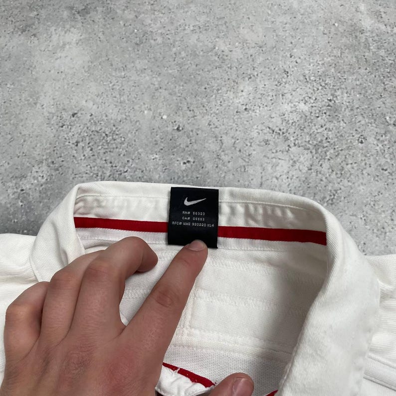 Pode incluir: Close-up de uma camisa polo branca com um detalhe de listra vermelha. A camisa tem uma etiqueta preta com o logotipo da Nike e texto. A camisa &eacute; feita de um tecido texturizado.