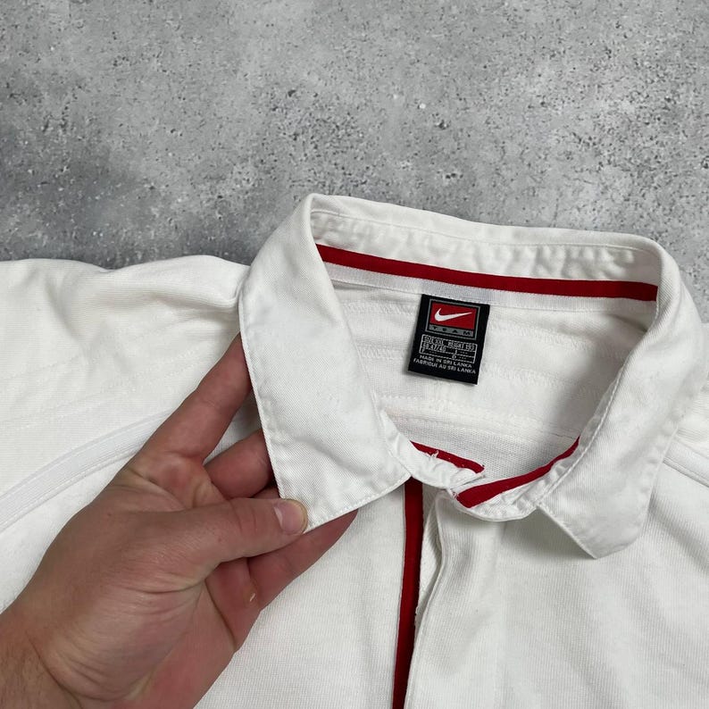 Pode incluir: Uma camisa branca com gola, acabamento vermelho e um logotipo da Nike Team. A camisa tem uma carcela abotoada com uma faixa vermelha no centro. A gola e o interior da gola s&atilde;o acabados a vermelho.