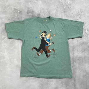 Pu&ograve; includere: T-shirt verde con un'illustrazione a cartoni animati di due figure che si abbracciano, circondate da stelle. Una figura indossa un abito e un cappello neri, l'altra una camicia blu e pantaloni marroni. La maglietta ha un girocollo.