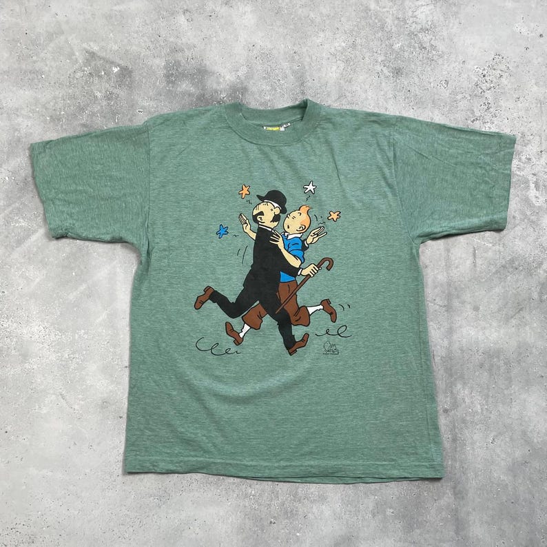 Pu&ograve; includere: T-shirt verde con un'illustrazione a cartoni animati di due figure che si abbracciano, circondate da stelle. Una figura indossa un abito e un cappello neri, l'altra una camicia blu e pantaloni marroni. La maglietta ha un girocollo.