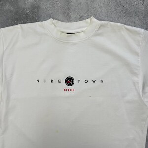 Peut inclure: Une tasse en c&eacute;ramique blanche avec une anse noire et le texte "But First, Coffee" en noir, en gras. La tasse est pos&eacute;e sur une surface blanche.