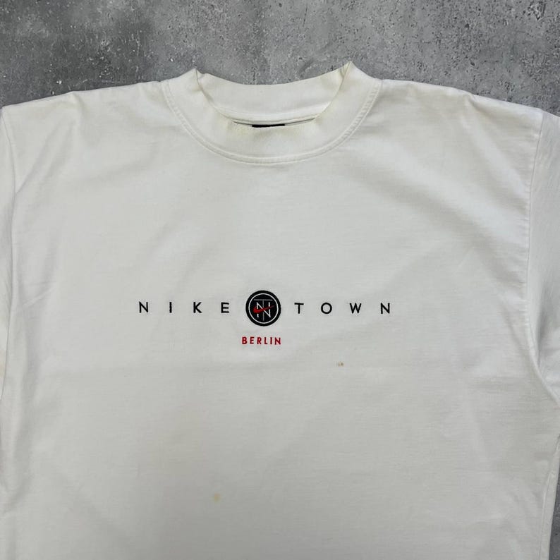 Peut inclure: Une tasse en c&eacute;ramique blanche avec une anse noire et le texte "But First, Coffee" en noir, en gras. La tasse est pos&eacute;e sur une surface blanche.