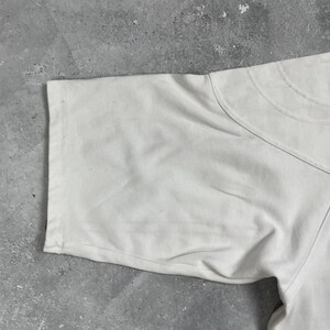 Pode incluir: Close-up de uma camiseta de manga curta de cor clara. O tecido parece ser macio, possivelmente algod&atilde;o. A manga &eacute; vis&iacute;vel, com costuras e decote redondo. A camisa &eacute; lisa e sem gr&aacute;ficos ou padr&otilde;es vis&iacute;veis.