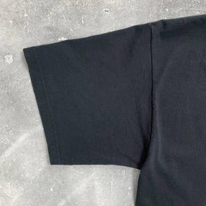 Op de afbeelding: Close-up van een zwarte t-shirtmouw. De mouw is gemaakt van een gladde, donkere stof en heeft een gestikte zoom. De afbeelding benadrukt de textuur en constructie van het kledingstuk.