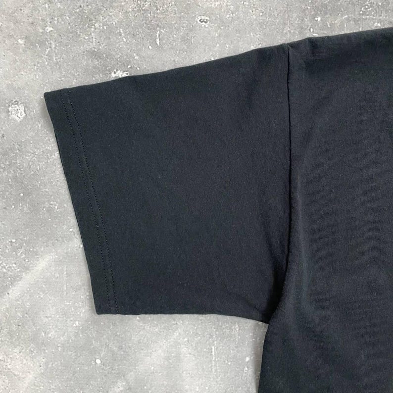 Op de afbeelding: Close-up van een zwarte t-shirtmouw. De mouw is gemaakt van een gladde, donkere stof en heeft een gestikte zoom. De afbeelding benadrukt de textuur en constructie van het kledingstuk.