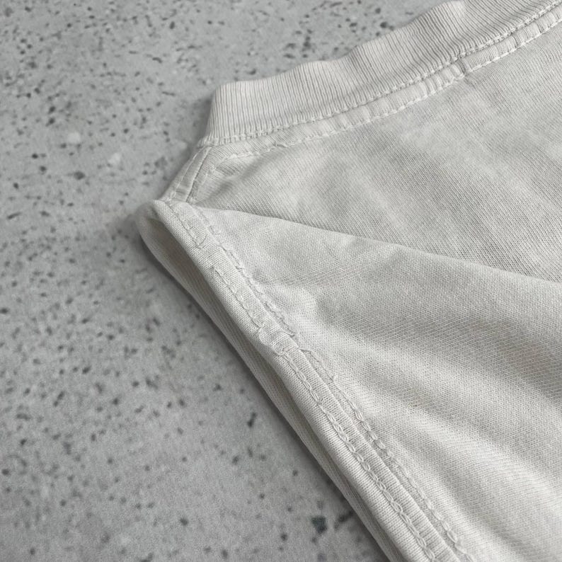 May include: Close-up of a light gray t-shirt. The fabric is soft and textured, with visible stitching along the seams of the neckline and shoulder. The shirt appears to be folded, with the edges of the fabric visible.