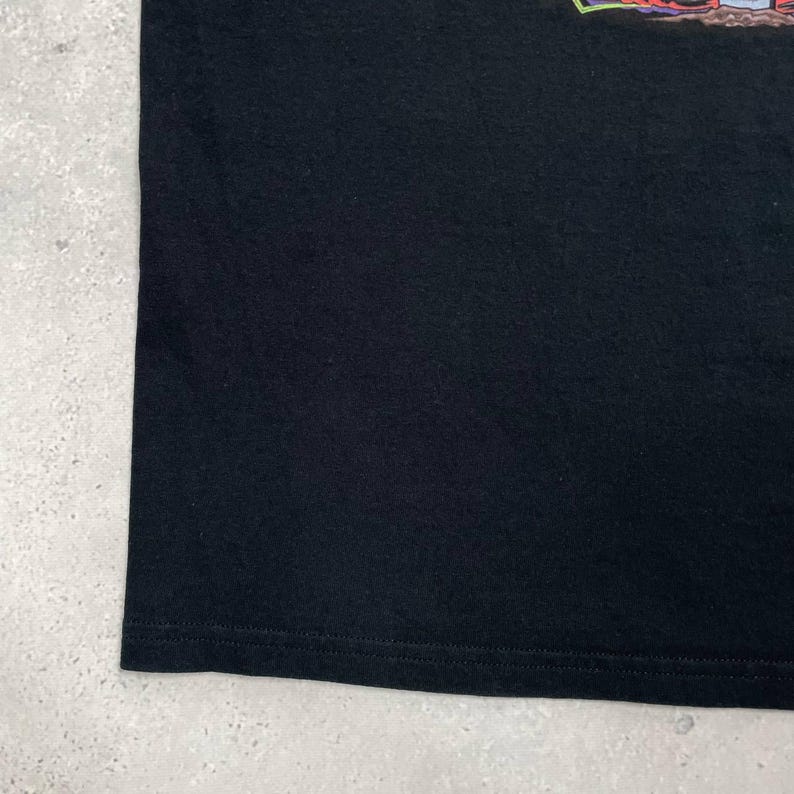 Op de afbeelding: Zwart T-shirt met een grafisch ontwerp aan de bovenkant. Het shirt is gemaakt van een zacht materiaal en heeft een gestikte zoom. De achtergrond is een lichtgrijze ondergrond.