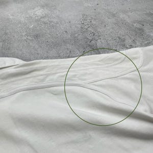 Pode incluir: Close-up de uma pe&ccedil;a de roupa clara, possivelmente uma camisa ou top, com costuras vis&iacute;veis e uma superf&iacute;cie texturizada. O tecido parece macio e ligeiramente enrugado.