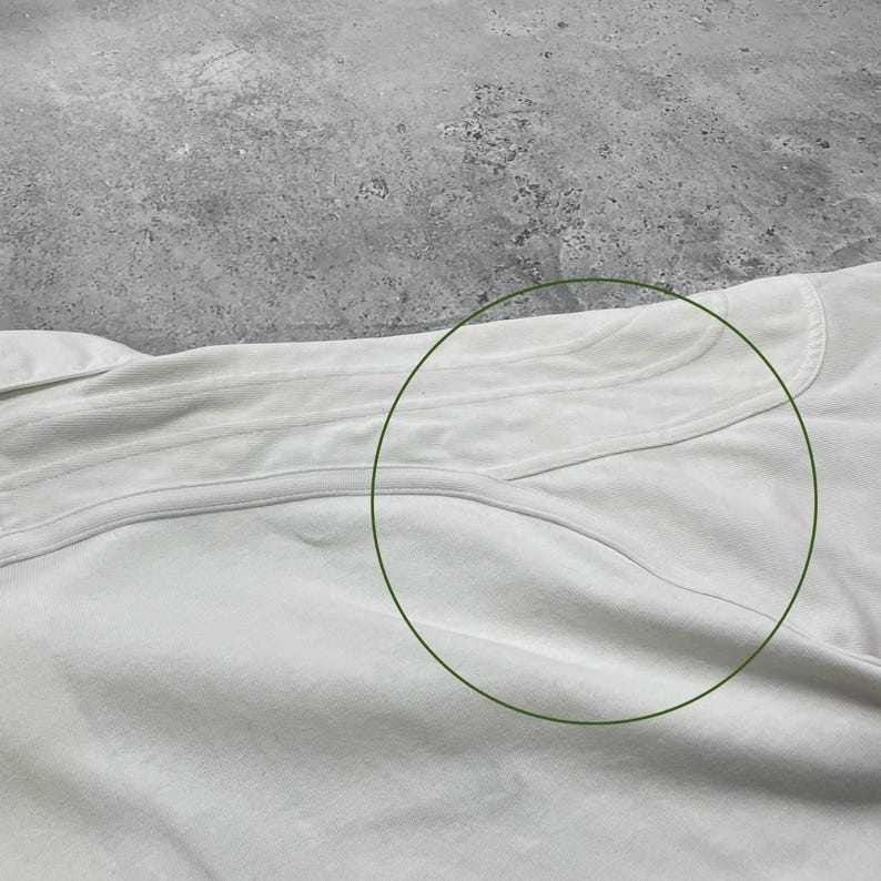 Pode incluir: Close-up de uma pe&ccedil;a de roupa clara, possivelmente uma camisa ou top, com costuras vis&iacute;veis e uma superf&iacute;cie texturizada. O tecido parece macio e ligeiramente enrugado.