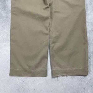 May include: Close-up of khaki-colored pants. The pants are made of a sturdy fabric and have a classic design. The hem of the pants is visible, with a slight fraying on one side. The pants are a neutral color, suitable for various outfits.