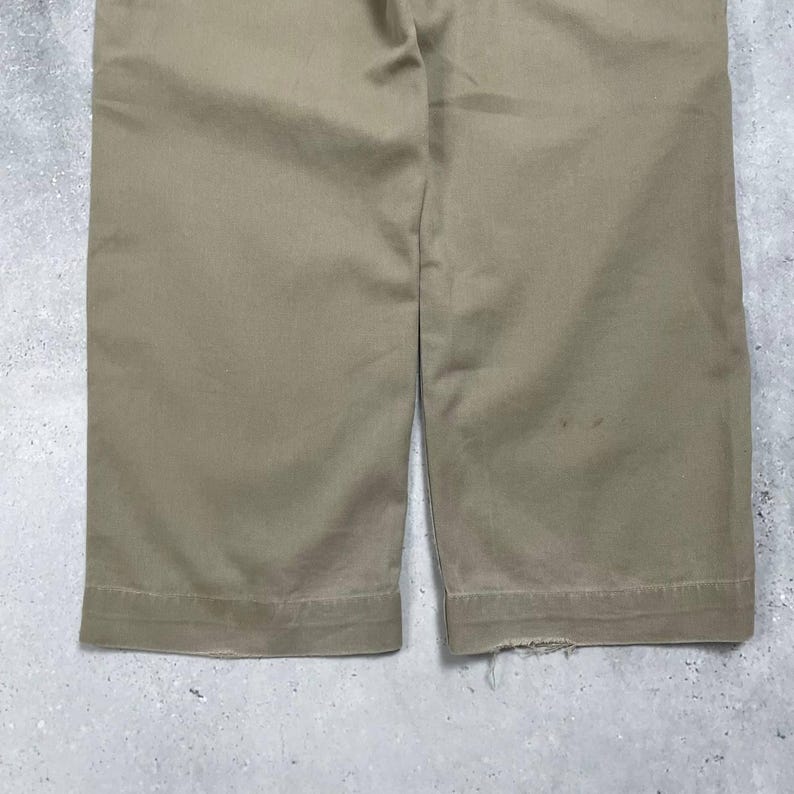 May include: Close-up of khaki-colored pants. The pants are made of a sturdy fabric and have a classic design. The hem of the pants is visible, with a slight fraying on one side. The pants are a neutral color, suitable for various outfits.