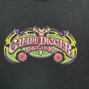 Op de afbeelding: Zwart T-shirt met een kleurrijke afbeelding. Het ontwerp toont de woorden "Grave Digger Race Team" in een gestileerd lettertype, met groene, paarse en rode accenten en sterren.