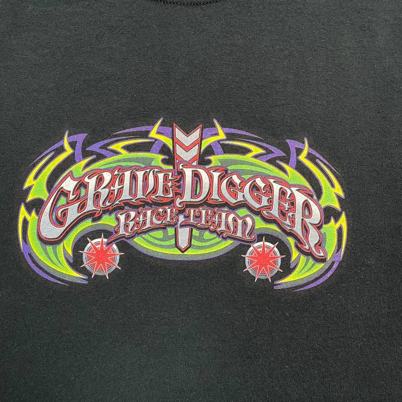 Op de afbeelding: Zwart T-shirt met een kleurrijke afbeelding. Het ontwerp toont de woorden "Grave Digger Race Team" in een gestileerd lettertype, met groene, paarse en rode accenten en sterren.