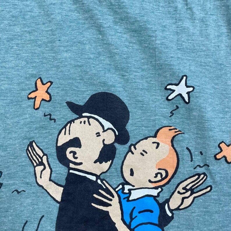 Pu&ograve; includere: T-shirt verde chiaro con un'illustrazione a cartoni animati di due personaggi. Un personaggio indossa un cappello e un abito neri, l'altro ha i capelli arancioni e una camicia blu. Stelle arancioni e bianche sono sparse intorno ai personaggi.