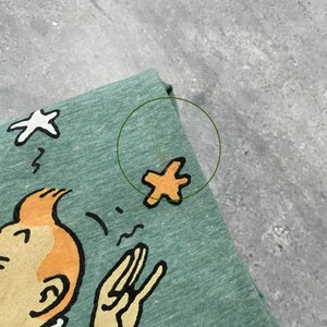 Pu&ograve; includere: Una maglietta verde chiaro con un'illustrazione di cartone animato. Il design include un personaggio con i capelli arancioni e un gesto della mano, insieme a elementi a forma di stella. La maglietta &egrave; realizzata in un tessuto morbido e strutturato.
