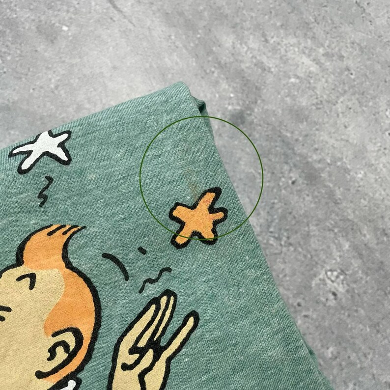 Pu&ograve; includere: Una maglietta verde chiaro con un'illustrazione di cartone animato. Il design include un personaggio con i capelli arancioni e un gesto della mano, insieme a elementi a forma di stella. La maglietta &egrave; realizzata in un tessuto morbido e strutturato.