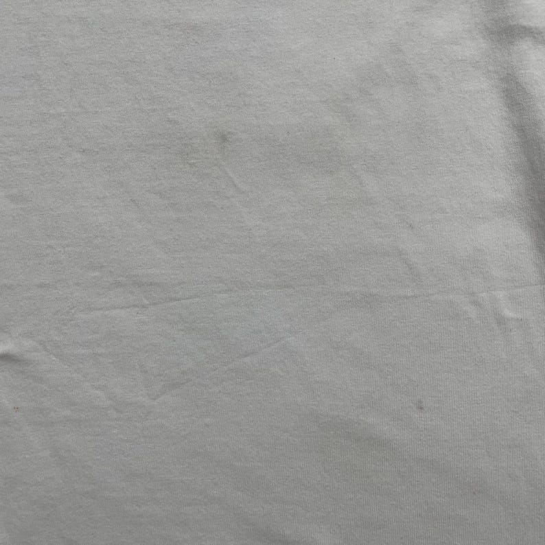 May include: Close-up of a white fabric with subtle variations in tone and texture. The material appears to be slightly wrinkled, with a few small, darker spots scattered across the surface. The overall impression is of a soft, plain textile.