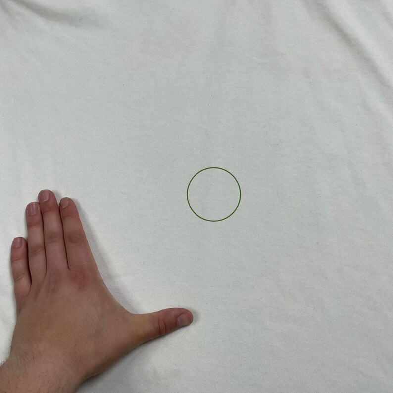 Pode incluir: Um tecido branco com um c&iacute;rculo verde desenhado. O tecido est&aacute; ligeiramente enrugado. Uma m&atilde;o &eacute; vis&iacute;vel no canto inferior esquerdo.