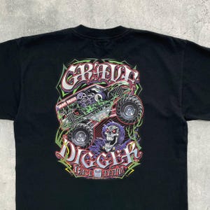 Op de afbeelding: Zwart t-shirt met een kleurrijke afbeelding van een monstertruck en de tekst "GRAVE DIGGER". Het ontwerp bevat een schedel en vlammen, met "RACE TEAM" onderaan. Het t-shirt is gemaakt van een zacht materiaal.