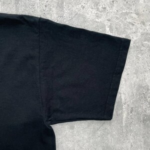 Op de afbeelding: Close-up van een zwarte t-shirtmouw. De mouw is gemaakt van een zachte, donkere stof en is langs de naden gestikt. De afbeelding is genomen vanuit een iets verhoogde hoek, die de textuur van het materiaal laat zien.