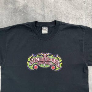 Op de afbeelding: Zwart T-shirt met een kleurrijk "Grave Digger" ontwerp. Het ontwerp bevat groene, paarse en gele accenten, met rode details. De tekst is in een gestileerd lettertype.