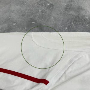 Pode incluir: Camiseta branca de manga comprida com uma faixa vermelha na parte inferior. A camisa tem gola redonda e &eacute; feita de um material macio, provavelmente algod&atilde;o. A imagem &eacute; um close-up, mostrando a textura do tecido.