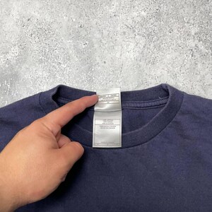 K&ouml;nnte beinhalten: Ein marineblaues T-Shirt mit Rundhalsausschnitt. Ein wei&szlig;es Etikett ist am Kragen befestigt. Das Etikett enth&auml;lt Text in Englisch und Franz&ouml;sisch. Das Shirt besteht aus 100 % Baumwolle.