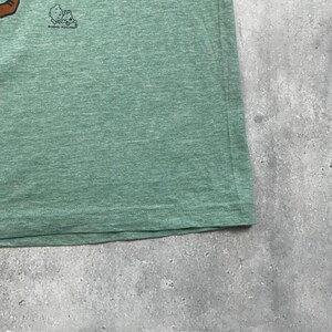 Pu&ograve; includere: Primo piano di una maglietta verde. Il tessuto ha una trama m&eacute;lange. Una piccola grafica e testo sono visibili nell'angolo in alto a sinistra. La maglietta sembra essere realizzata in un materiale morbido e confortevole.