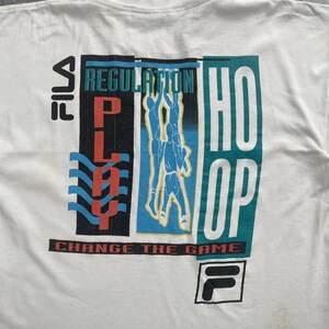 May include: White t-shirt with a colorful graphic design. The design includes the words "PLAY", "REGULATION", and "HOOP" in bold letters. The shirt also features a basketball player illustration and the FILA logo. The text "CHANGE THE GAME" is printed at the bottom.