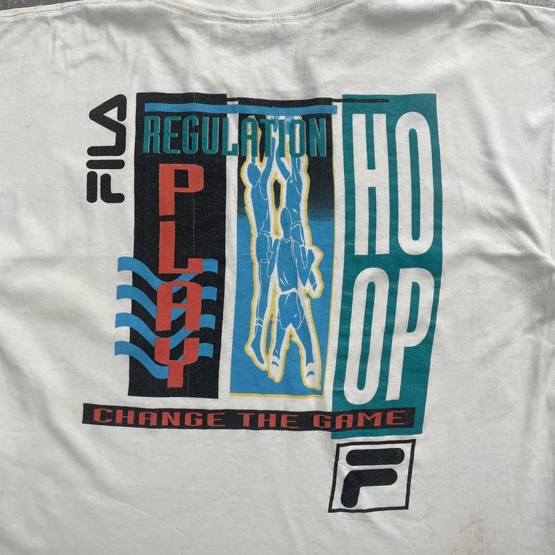May include: White t-shirt with a colorful graphic design. The design includes the words "PLAY", "REGULATION", and "HOOP" in bold letters. The shirt also features a basketball player illustration and the FILA logo. The text "CHANGE THE GAME" is printed at the bottom.