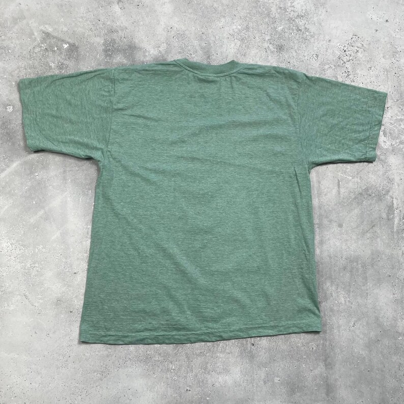 Pu&ograve; includere: Una maglietta verde tinta unita a maniche corte. La maglietta &egrave; realizzata in un tessuto morbido e strutturato e ha uno scollo tondo. La maglietta &egrave; stesa su una superficie grigia.