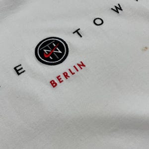 Peut inclure: Sweat-shirt blanc avec texte et logo brod&eacute;s. Le mot "BERLIN" est brod&eacute; en rouge, et le logo est un cercle noir avec un swoosh rouge et les lettres "TN" &agrave; l'int&eacute;rieur. Le mot "TOWN" est brod&eacute; en noir.