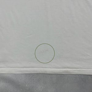 Pode incluir: Uma camisa branca em close-up com uma pequena mancha. A bainha inferior da camisa &eacute; vis&iacute;vel, juntamente com uma superf&iacute;cie cinza abaixo. Um c&iacute;rculo verde envolve a mancha.