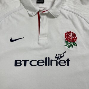 Pode incluir: Camisa de rugby branca com um emblema de rosa vermelha e o texto "BT cellnet" em azul marinho. Apresenta um logotipo Nike swoosh azul marinho. A camisa tem gola e uma carcela com acabamento vermelho.