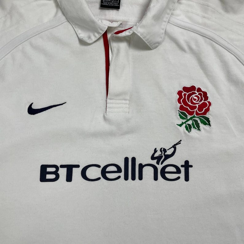 Pode incluir: Camisa de rugby branca com um emblema de rosa vermelha e o texto "BT cellnet" em azul marinho. Apresenta um logotipo Nike swoosh azul marinho. A camisa tem gola e uma carcela com acabamento vermelho.