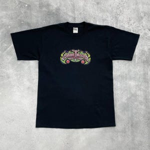 Op de afbeelding: Zwart t-shirt met een kleurrijke afbeelding. De afbeelding toont de tekst "Grave Digger" in een gestileerd lettertype, met groene, gele en paarse accenten. Twee rode accenten bevinden zich aan de onderkant van de afbeelding. Het shirt is gemaakt van zacht materiaal.