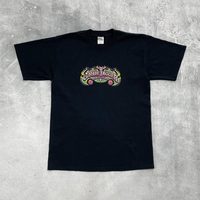 Op de afbeelding: Zwart t-shirt met een kleurrijke afbeelding. De afbeelding toont de tekst "Grave Digger" in een gestileerd lettertype, met groene, gele en paarse accenten. Twee rode accenten bevinden zich aan de onderkant van de afbeelding. Het shirt is gemaakt van zacht materiaal.