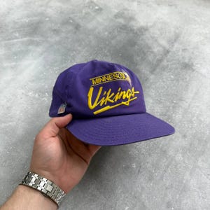 以下が含まれることがあります： 紫色の野球帽。前面に黄色の筆記体で「MINNESOTA Vikings」の文字が書かれています。NFLのロゴが側面に表示されています。