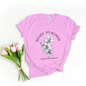 May include: Pink t-shirt with a black and white graphic of lilies and the text "Ready to Bloom" and "Bloom with Strength".