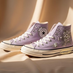 Può includere: Un paio di scarpe da ginnastica Converse alte lilla con lacci bianchi e suola color crema. Le scarpe da ginnastica presentano ricami floreali e il logo Converse All Star. Il testo "insightfull ives STORE" è stampato sul lato.