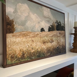 May include: A framed painting of a landscape with a field of tall grass in the foreground, a line of trees in the middle ground, and a cloudy sky in the background. The painting is in a brown wooden frame.