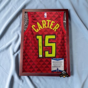 May include: Framed basketball jersey with the name "CARTER" and the number "15" in yellow. The jersey is red with a geometric pattern. A Beckett Certificate is visible. The frame is a dark brown color.