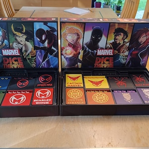 Peut inclure: Deux boîtes de jeu Marvel Dice Throne ouvertes sur une table en bois. Chaque boîte présente des illustrations de personnages et contient des cartes et des dés. Les boîtes présentent des personnages comme Thor, Spider-Man et Black Panther. Les boîtes de jeu sont noires avec des graphismes colorés.